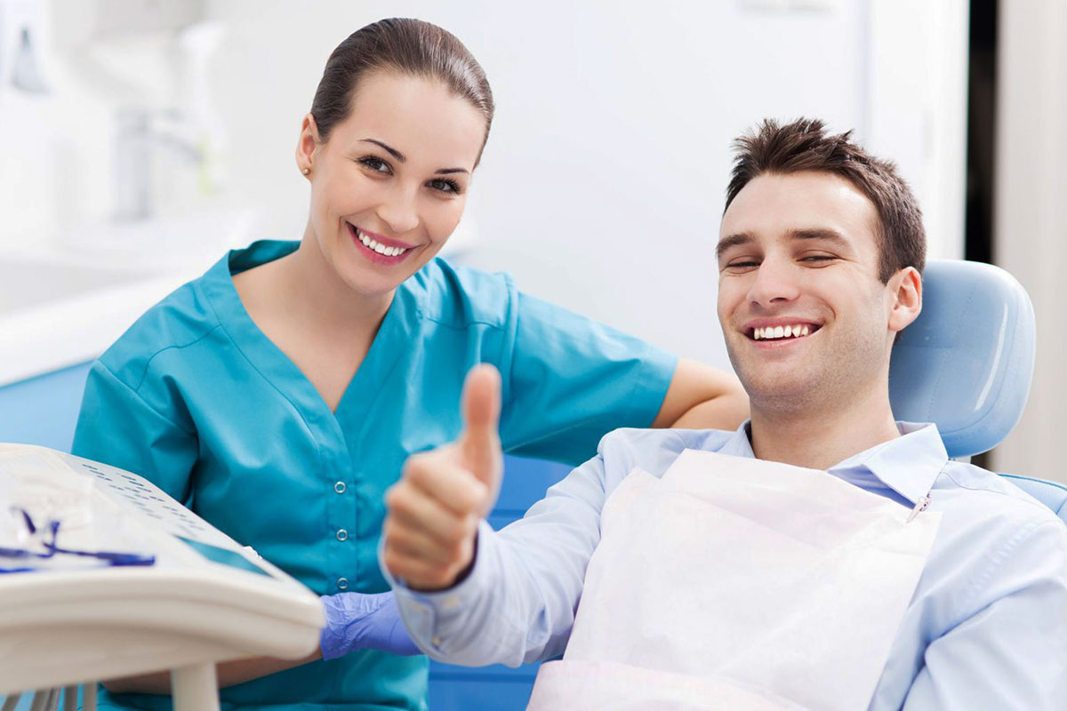 Smiling dental professional in scrubs with satisfied male patient giving thumbs up in dentist office.