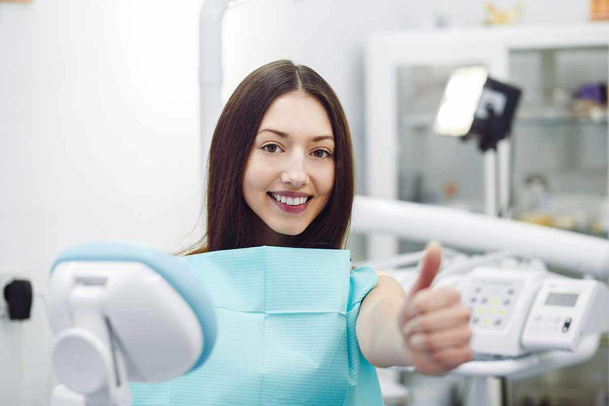 Young woman in dental bib giving thumbs up in modern dental office treatment room.