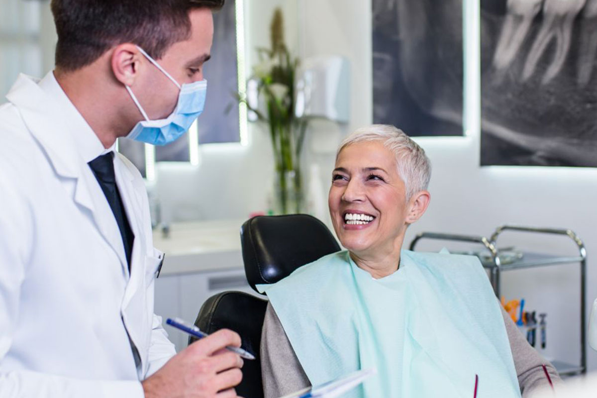 Smiling senior patient in dental bib speaking with masked dentist in modern office setting.