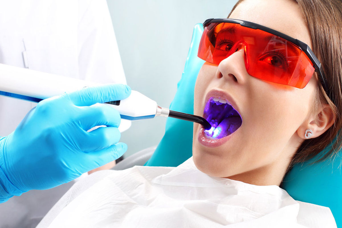 Patient receiving dental treatment with UV curing light while wearing protective red glasses at dental office.