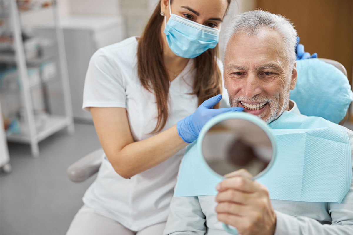 Senior man examines teeth in mirror while female dentist in mask assists in dental office.