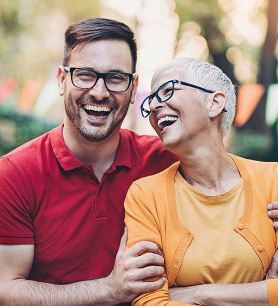 Young man and older woman smiling together outdoors wearing glasses, warm clothing.