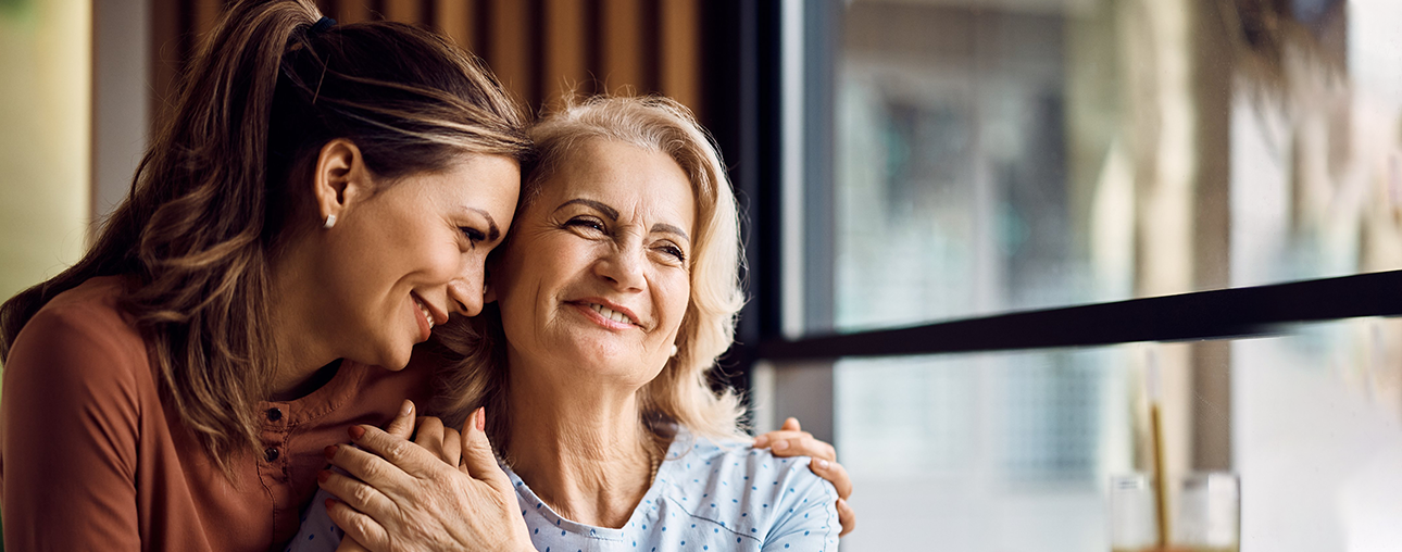 Adult daughter embracing smiling senior mother by bright window in warm home setting.