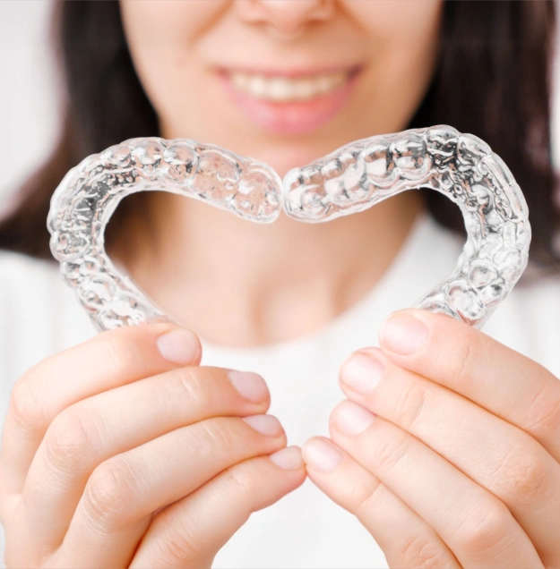 Woman holding clear dental retainer shaped like a heart, smiling in background.