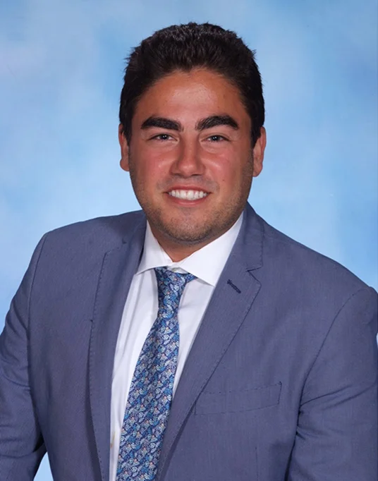 Professional headshot of man in blue suit and patterned tie against light blue background.